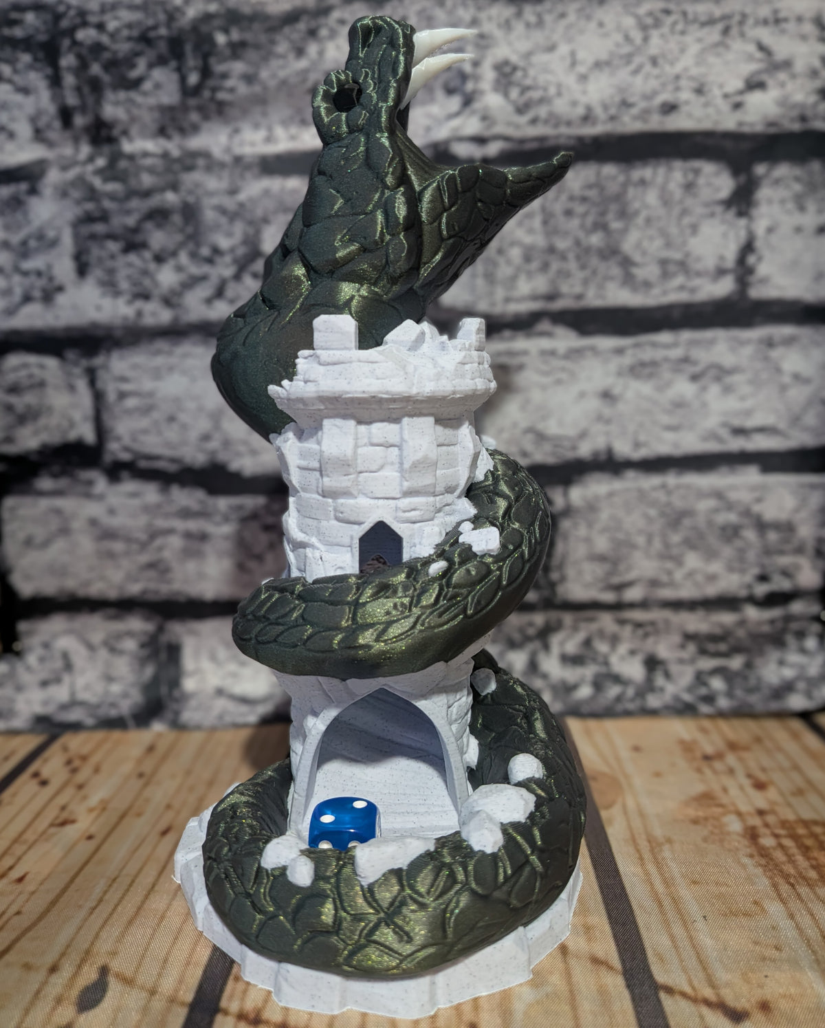A fantasy tabletop miniature featuring a dark green dragon coiled around a white stone tower, with a small blue dice piece in an archway at the base, set against a faux brick wall and wooden surface.