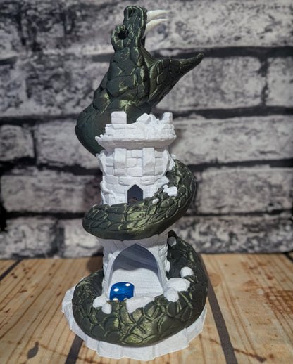 A fantasy tabletop miniature featuring a dark green dragon coiled around a white stone tower, with a small blue dice piece in an archway at the base, set against a faux brick wall and wooden surface.