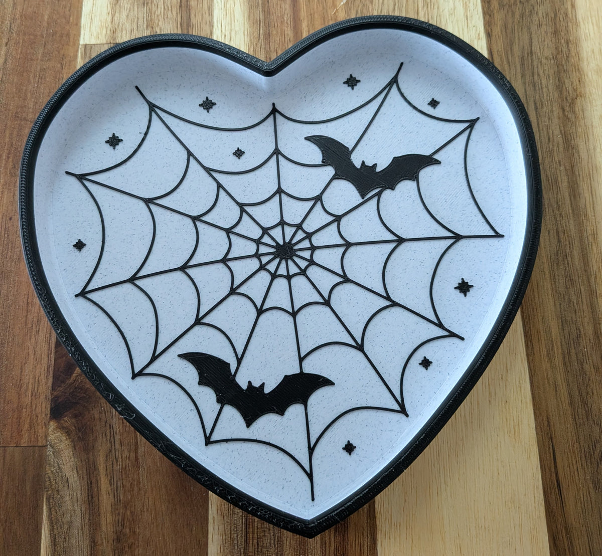 A heart-shaped Halloween decorative tray with a black spiderweb design, black bat silhouettes, and small star accents on a white background, set on a wooden surface.
