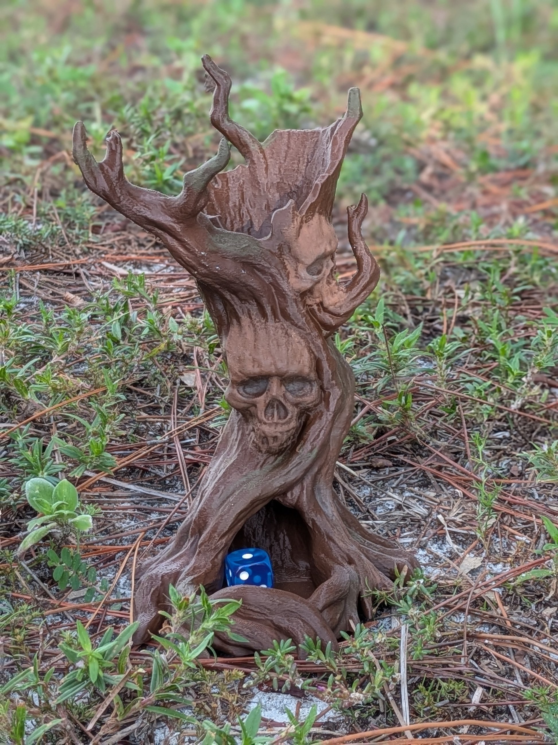 A small carved wooden tree stump figurine with skull faces and antler-like branches, placed outdoors in grass and pine needles. A small blue die rests inside the hollow base of the stump.