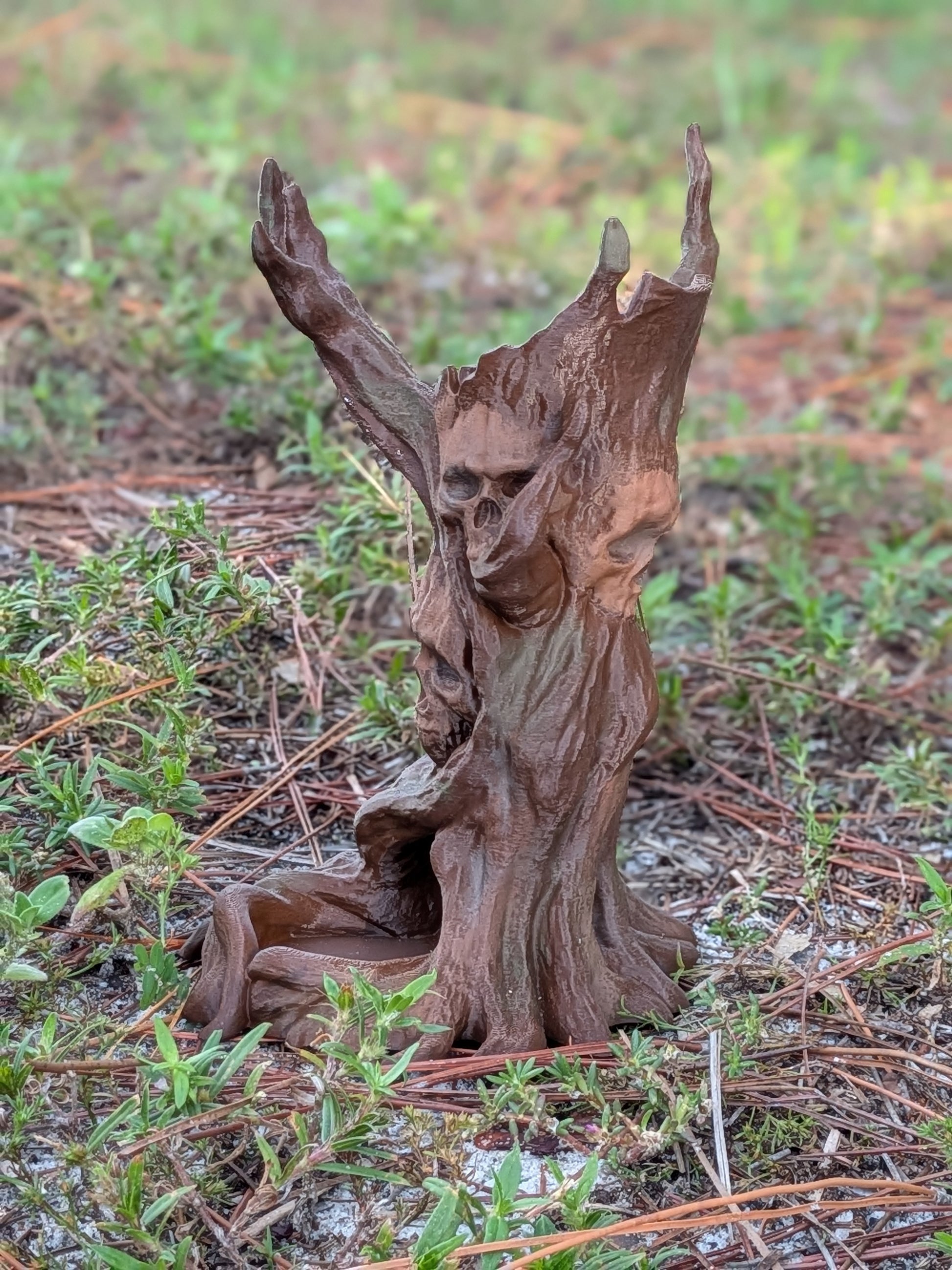 A small sculpted tree stump figurine with a skull face and raised branches, placed outdoors on grass and pine needles.