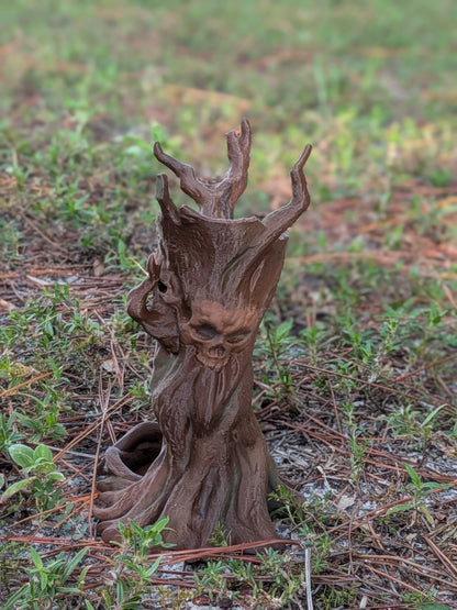 A small brown tree stump figurine with a carved face and branching antler-like tops, placed outdoors on grass and pine needles.
