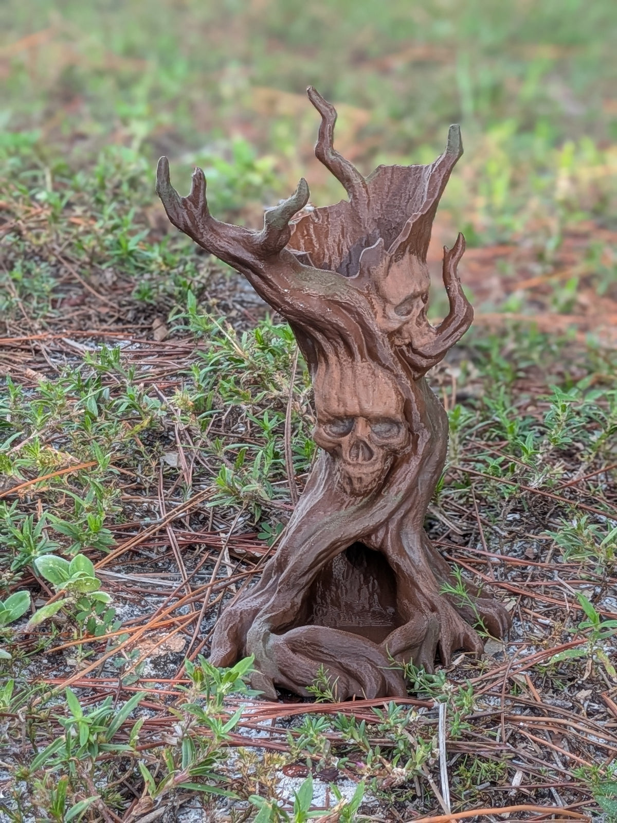 A small decorative tree stump sculpture with a carved skull face and branching antler-like tops, placed outdoors on grass and pine needles.
