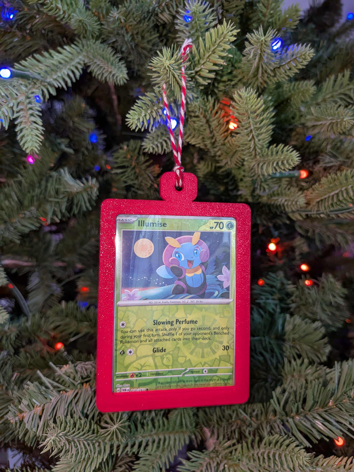 A Pokémon TCG ornament shaped like a red-bordered Illume card hanging from a green Christmas tree with colorful string lights.