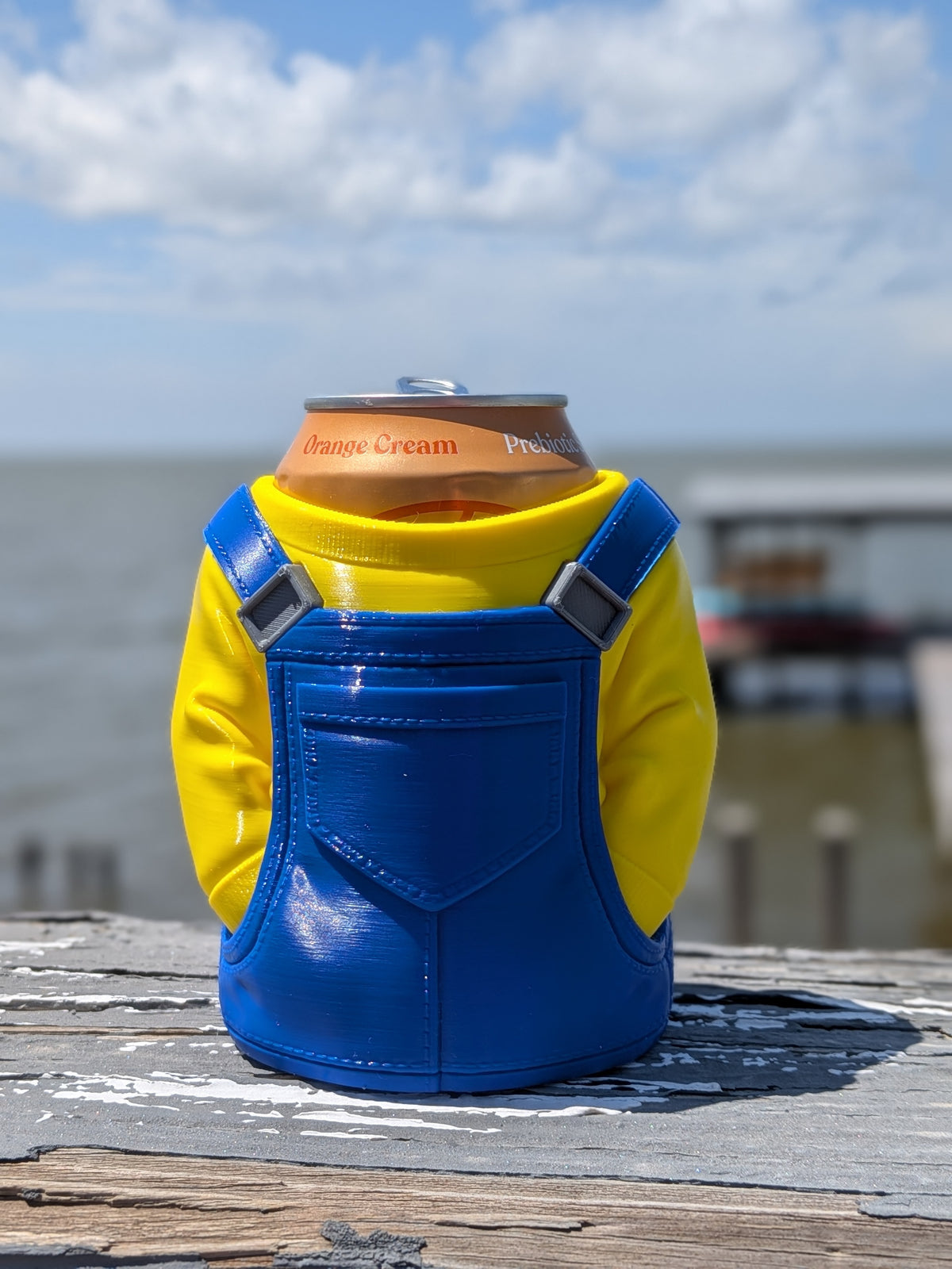 3D-printed can holder shaped like a yellow character in blue overalls, holding a soda can on a dock railing