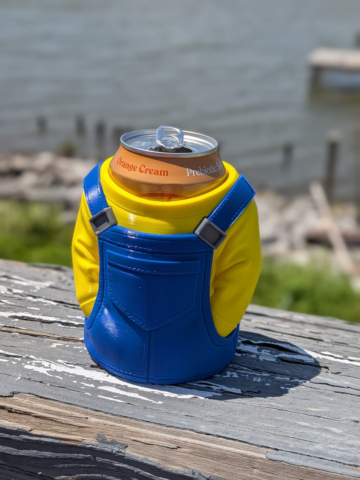 Yellow overalls character can holder with a soda can, front view outdoors near water